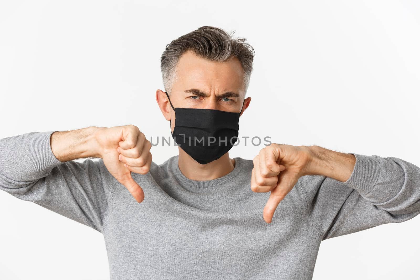 Concept of covid-19, social distancing and quarantine. Close-up of angry and disappointed middle-aged man in black medical mask, frowning upset and showing thumbs-down.