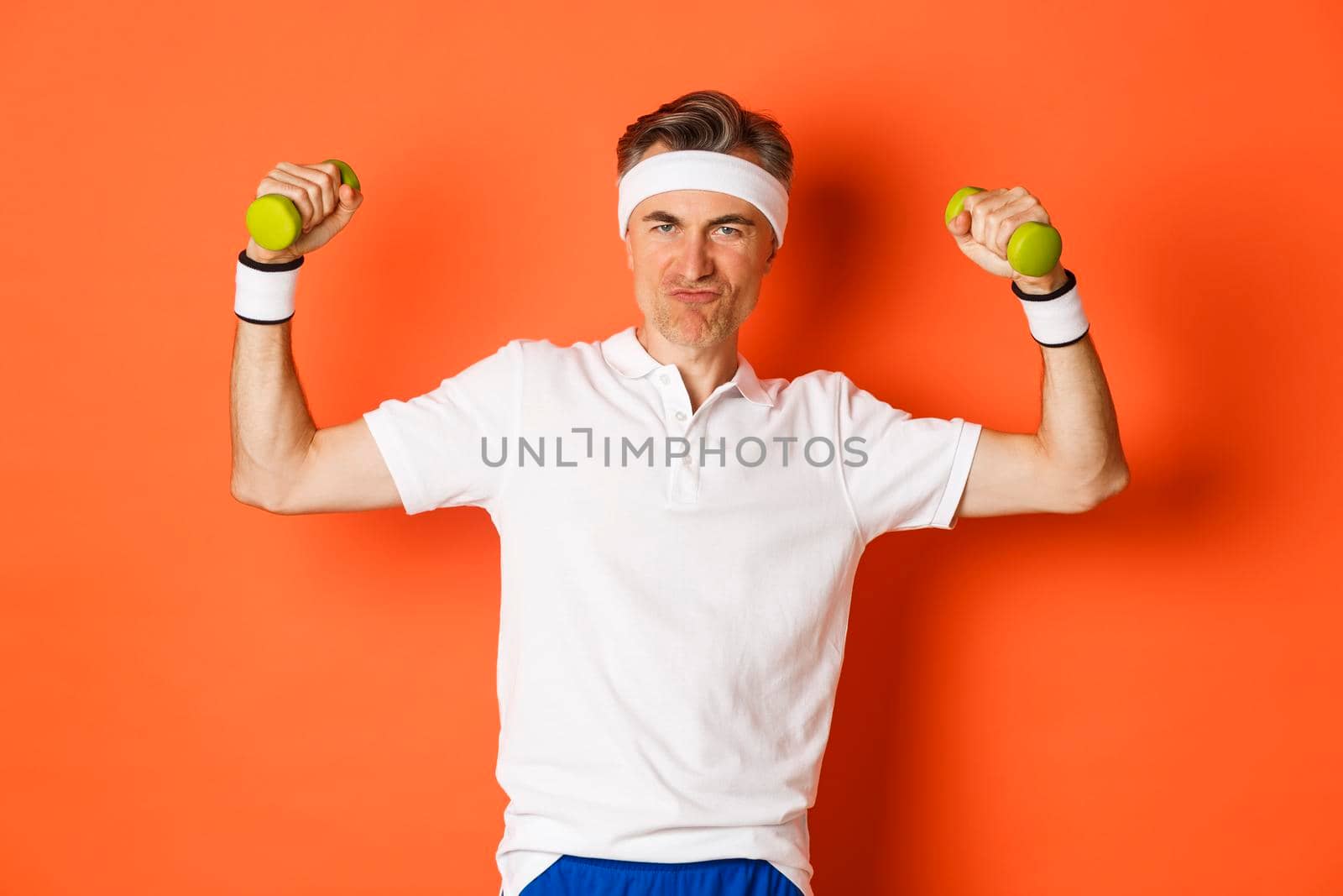 Portrait of cheeky middle-aged fitness guy, doing sports against orange background, flex biceps and holding dumbbells.