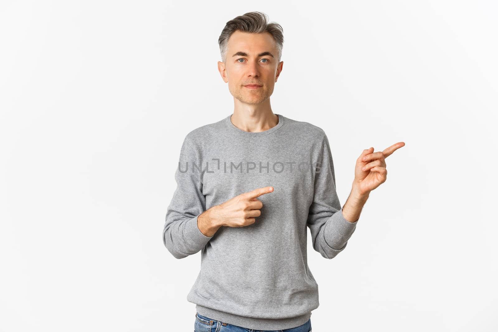 Portrait of attractive middle-aged man in grey sweater, pointing fingers right and looking serious, showing an advertisement, standing over white background.