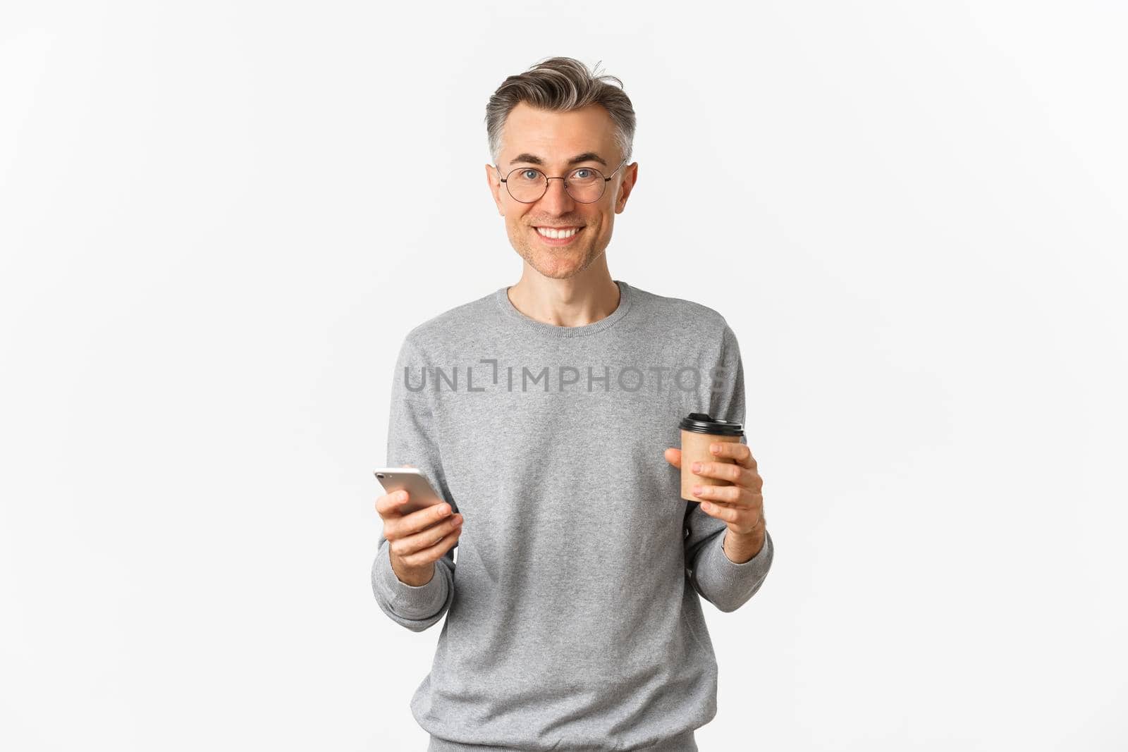 Handsome middle-aged man in grey sweater and glasses, smiling happy, drinking coffee and using mobile phone, standing over white background.