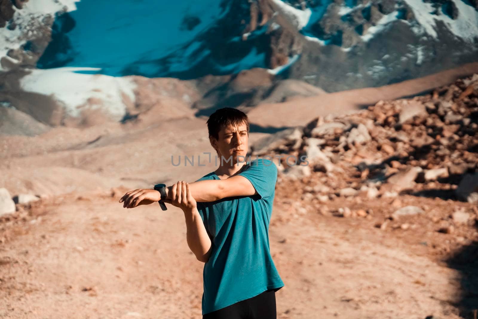 A young man does a warm-up before a trail and canyon race in the snow-capped mountains. The runner trains outdoor, does exercises in beautiful area.