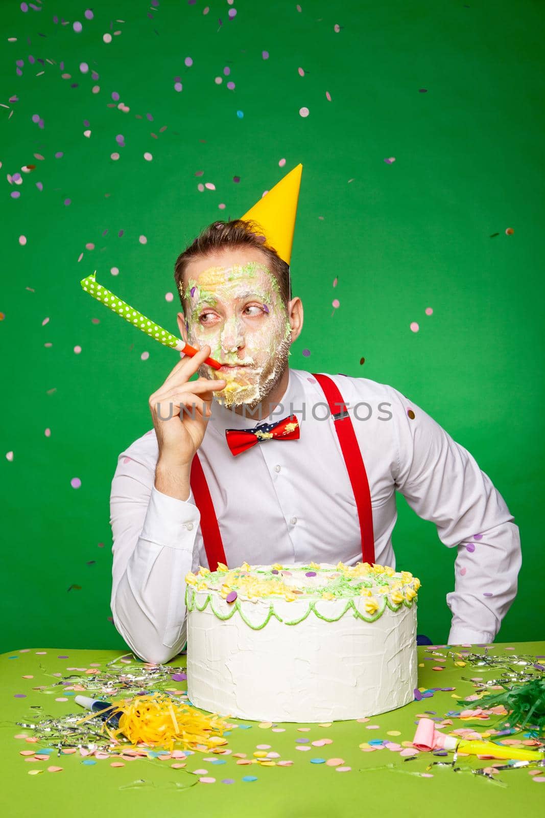 Male with face smeared in birthday cake sitting at table and blowing noisemaker during party in studio on green background