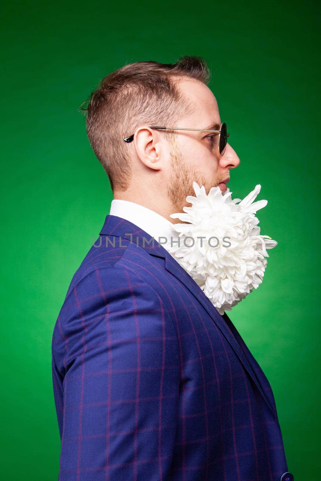 Self assured male in elegant suit and with tender white flowers in beard looking at camera on green background in studio