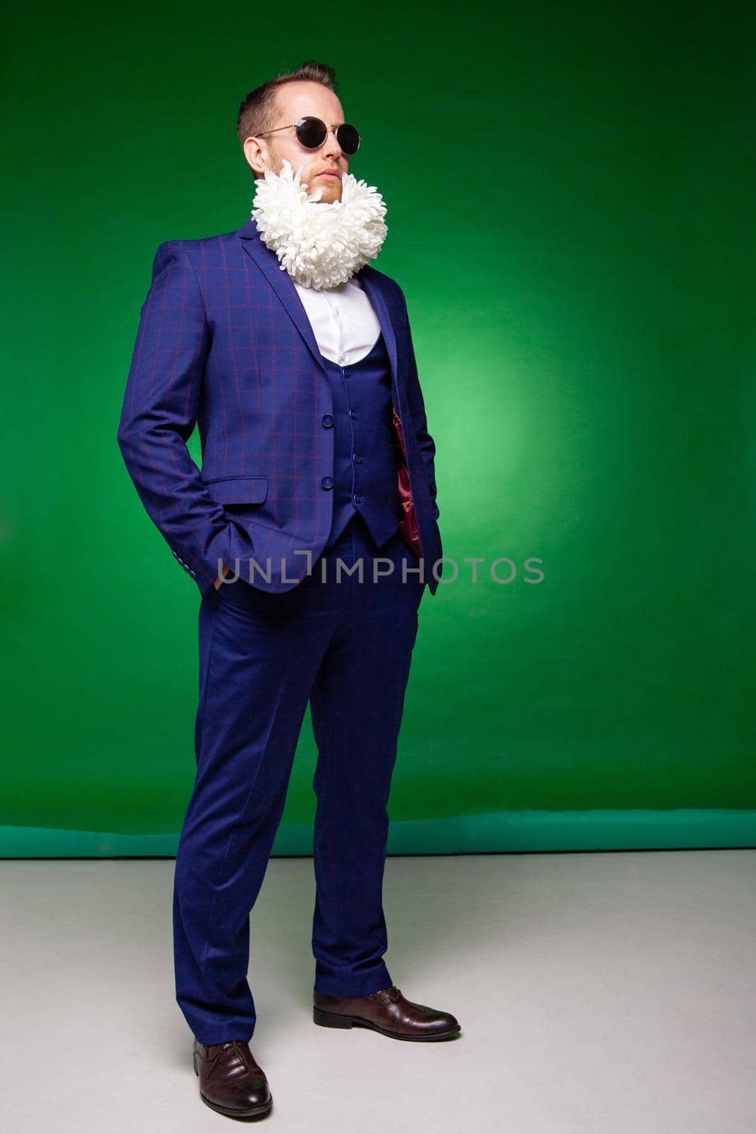 Confident male in elegant suit and sunglasses standing with white flowers in beard in studio on green background