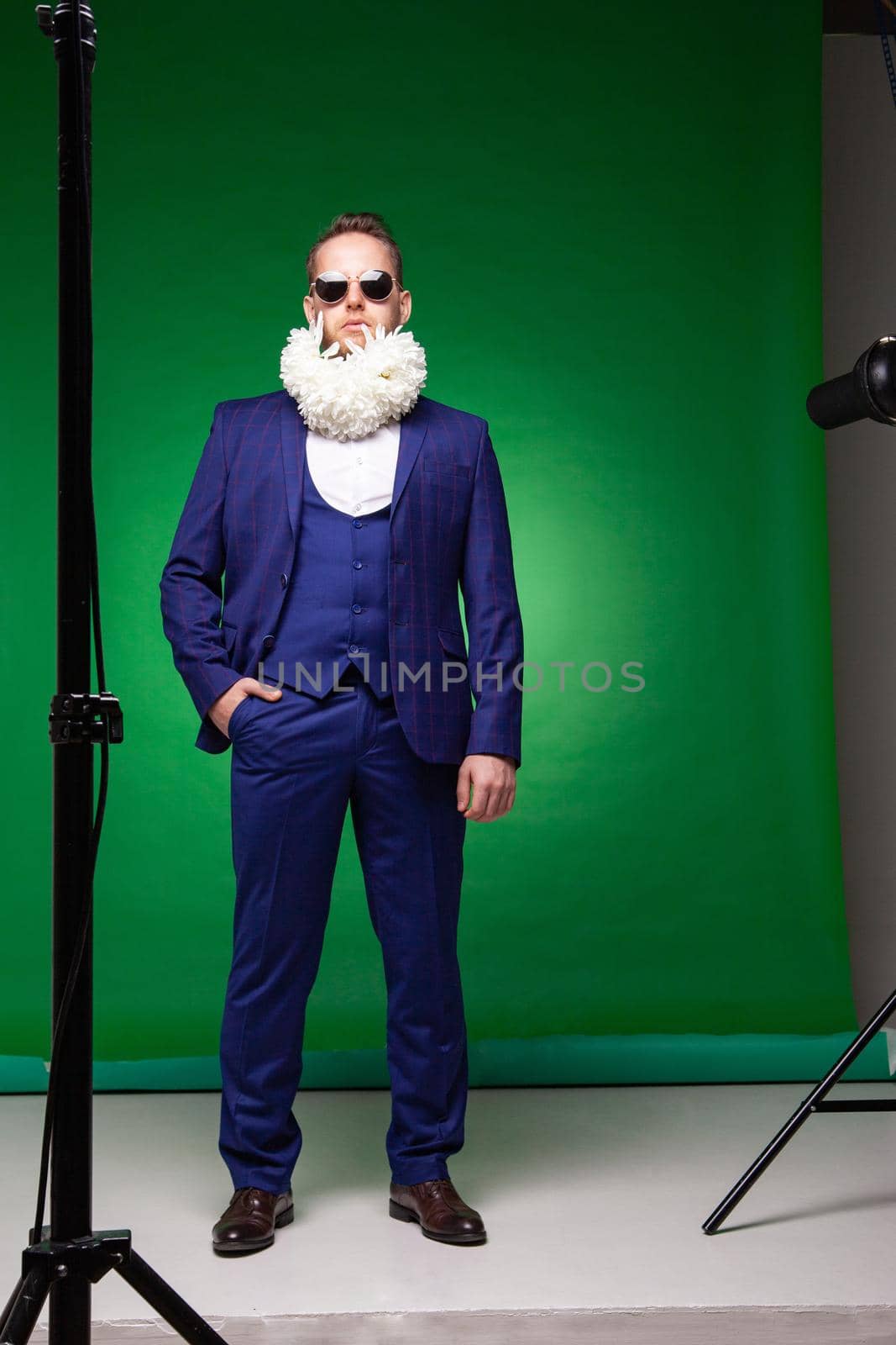 Confident male in elegant suit and sunglasses standing with white flowers in beard in studio on green background