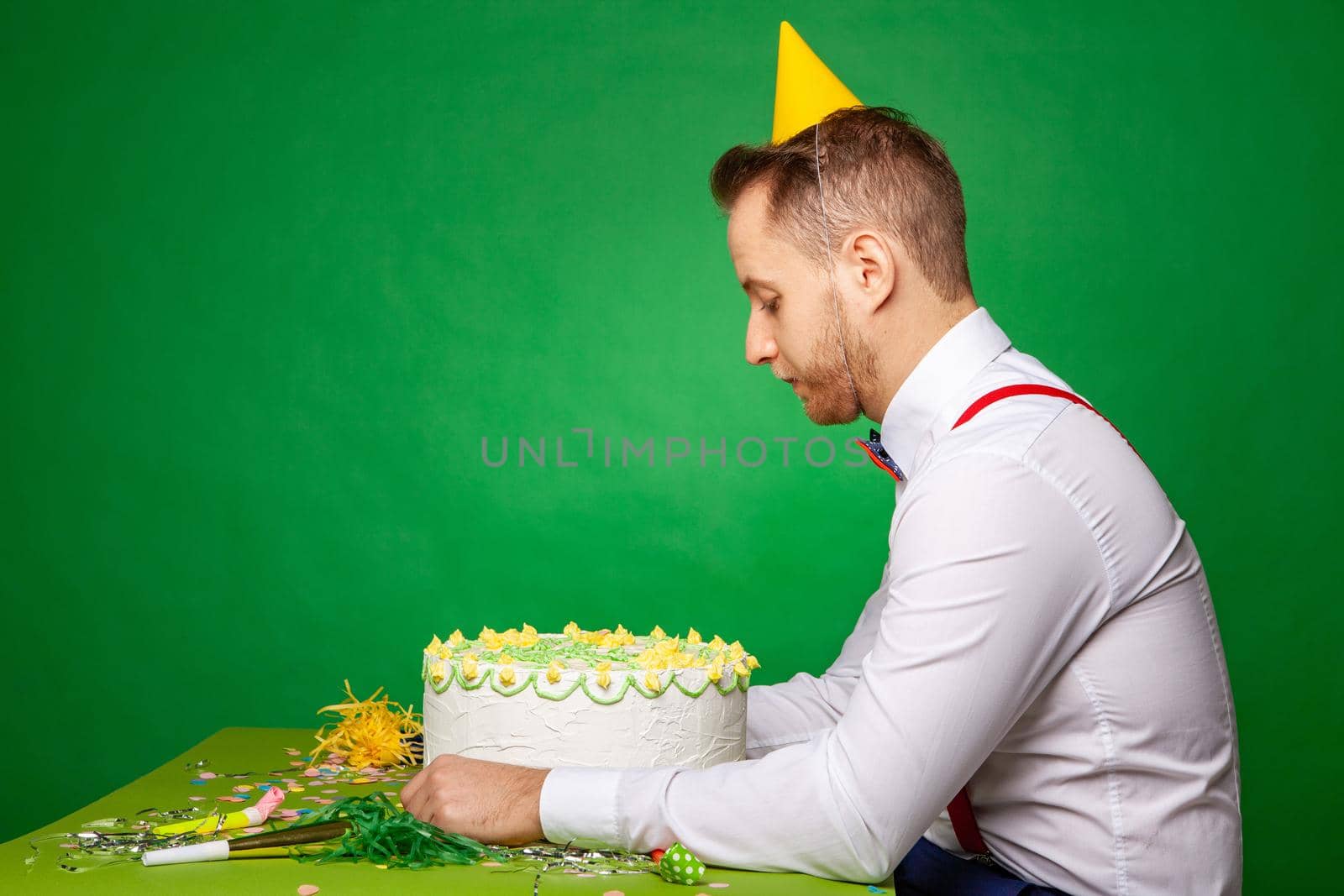 Upset male in party hat sitting at table with birthday cake and leaning on hand on green background in studio