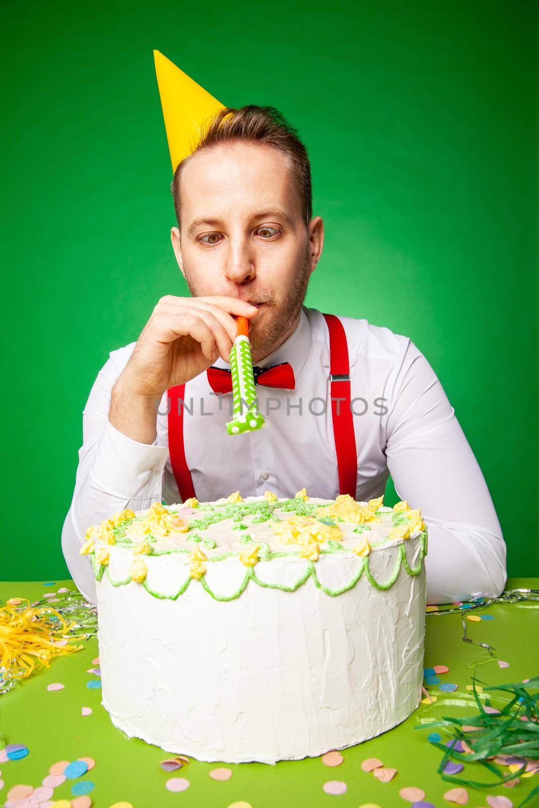 Man in party hat sitting at table with sweet birthday cake and blowing noisemaker on green backwound in studio