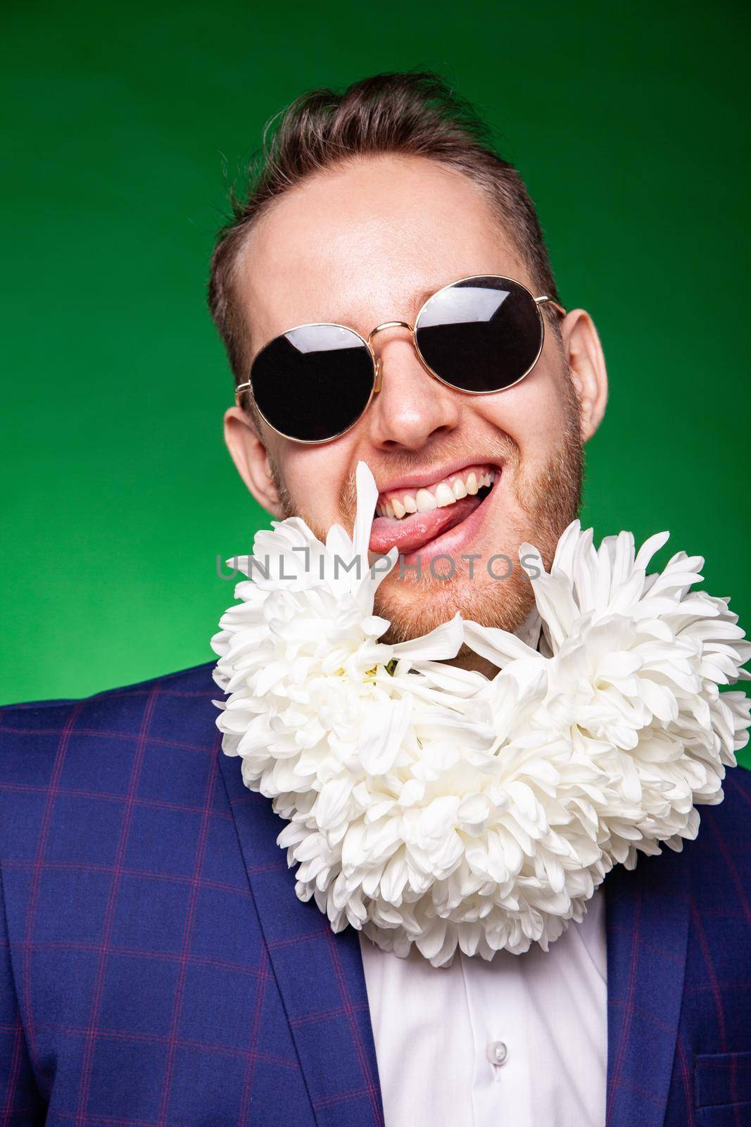 Funny male in suit and sunglasses licking petal of flowers while having fun in studio on green background and looking at camera
