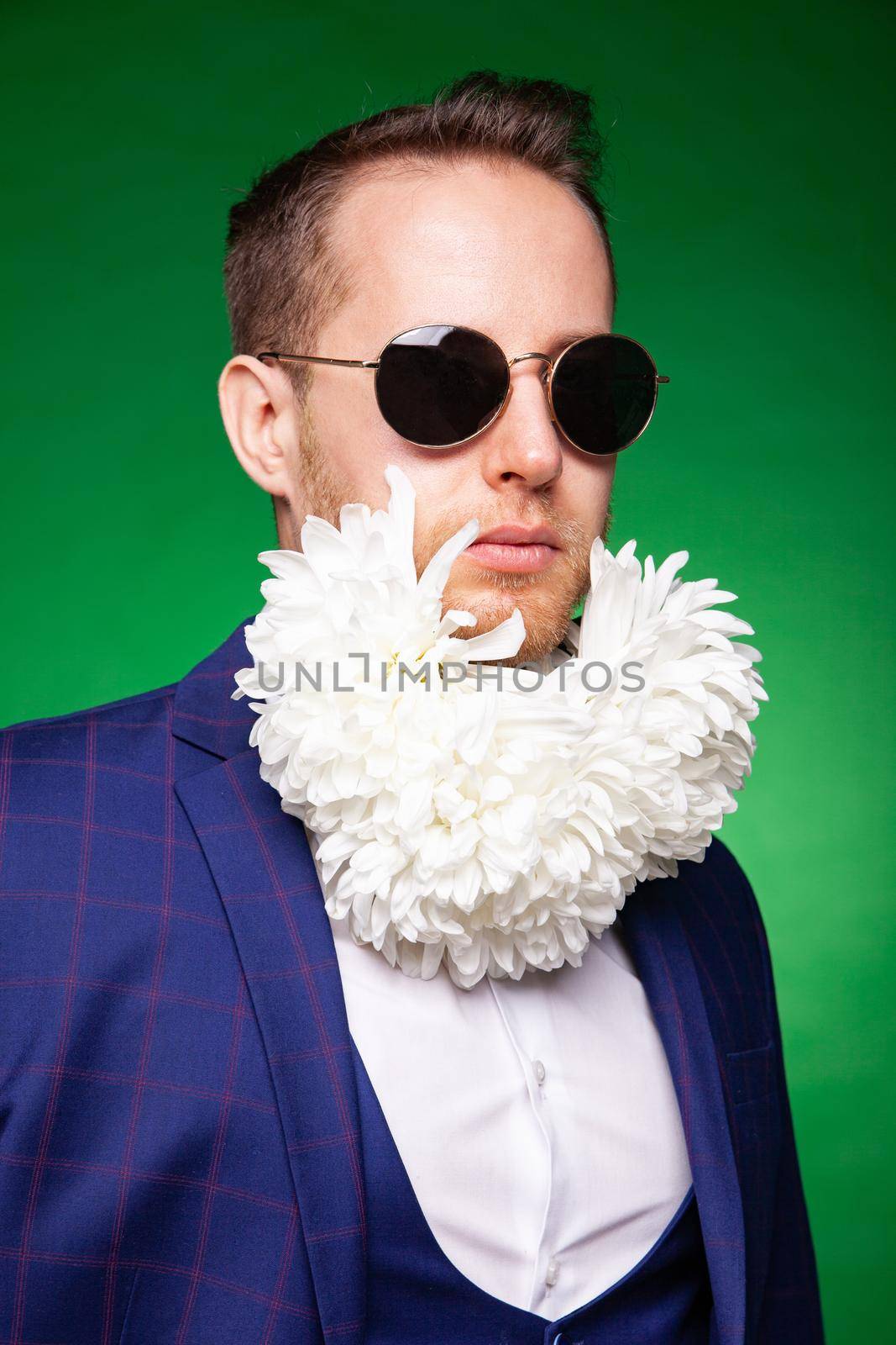 Self assured male in elegant suit and with tender white flowers in beard looking at camera on green background in studio