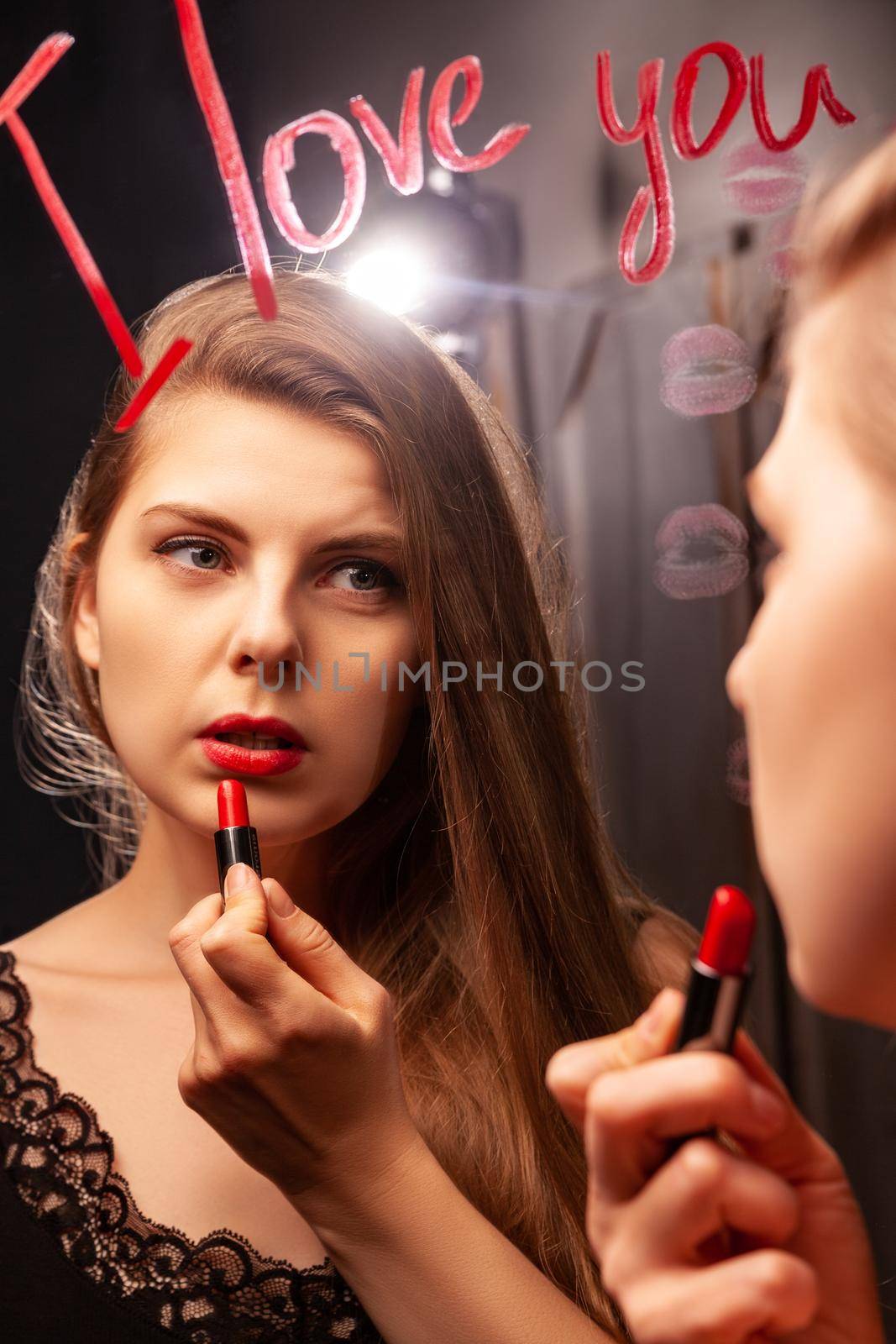 Lovely female looking in mirror with inscription I Love You and applying red lipstick while doing makeup