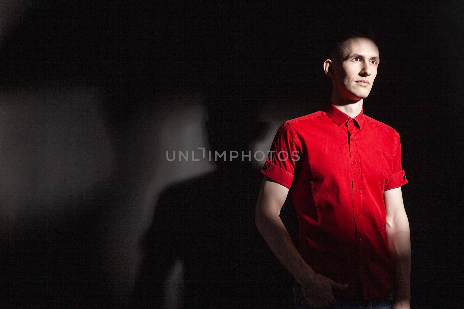 Serious confident young male in bright red shirt looking away while standing under light in dark studio with black wall and shadow