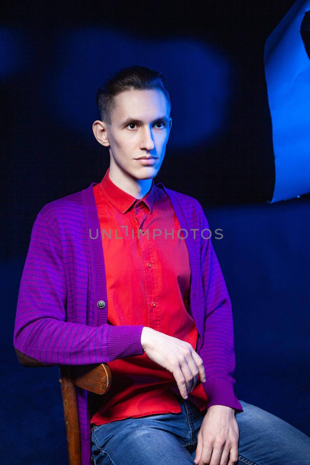 Young male in bright red shirt and purple blouse looking away while sitting on chair in dark studio with black and blue wall and shadow