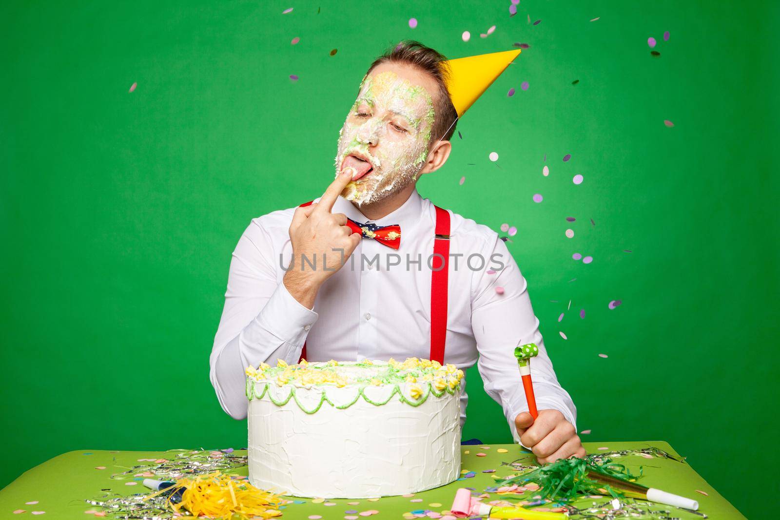 Male in party hat sitting at table and tasting sweet delicious birthday cake while looking up on green background in studio