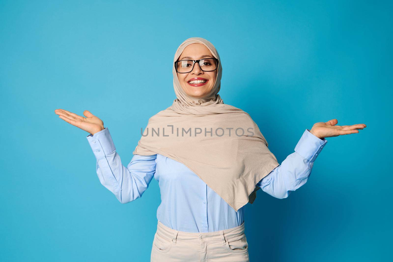 Smiling woman with covered head in hijab posing with outstretched arms and palms up, presenting on blue background with space for text