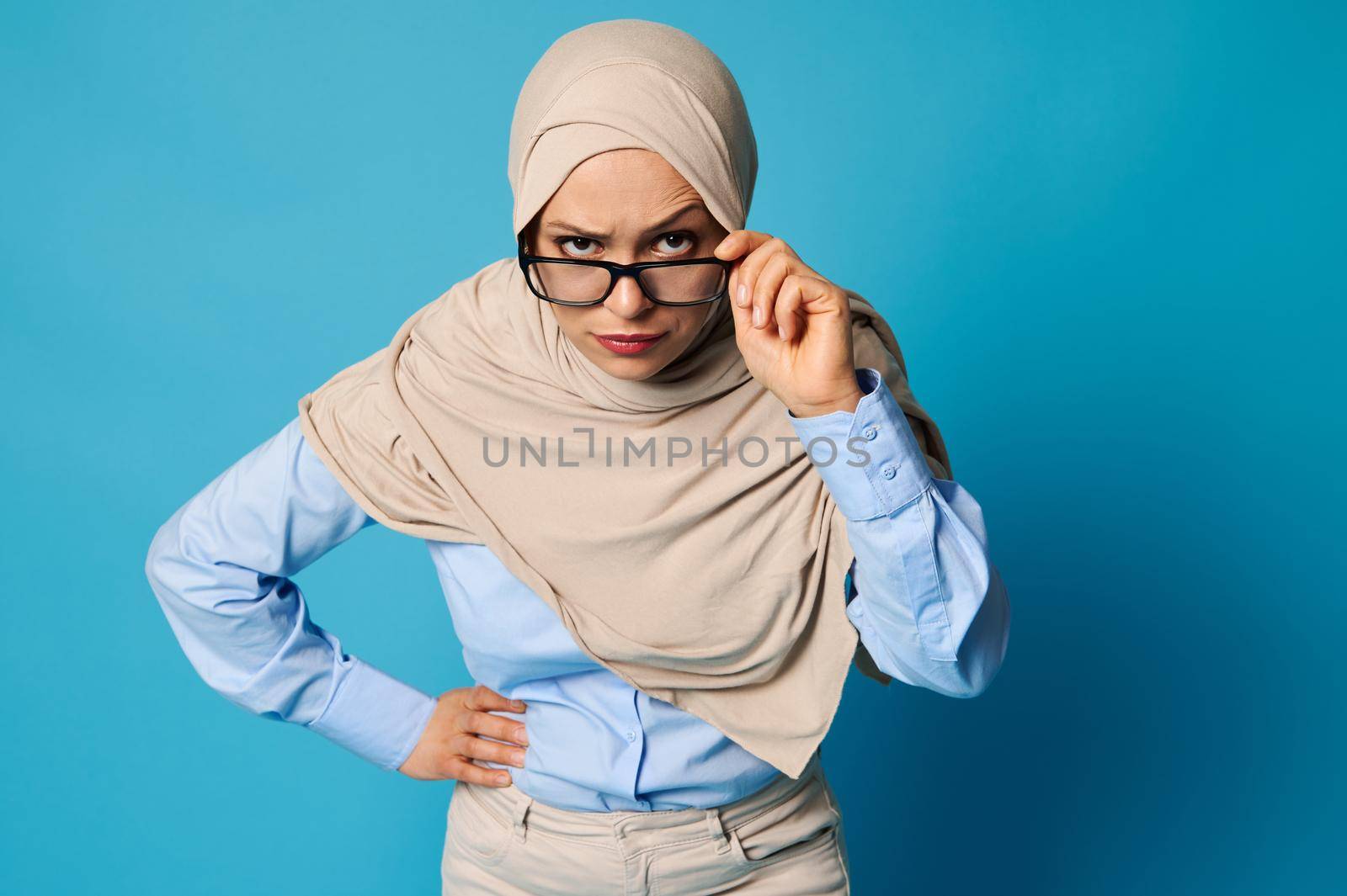 Portrait of a Muslim woman in hijab holding eyeglasses by the temple and looking carefully through them with raised eyebrow by artgf