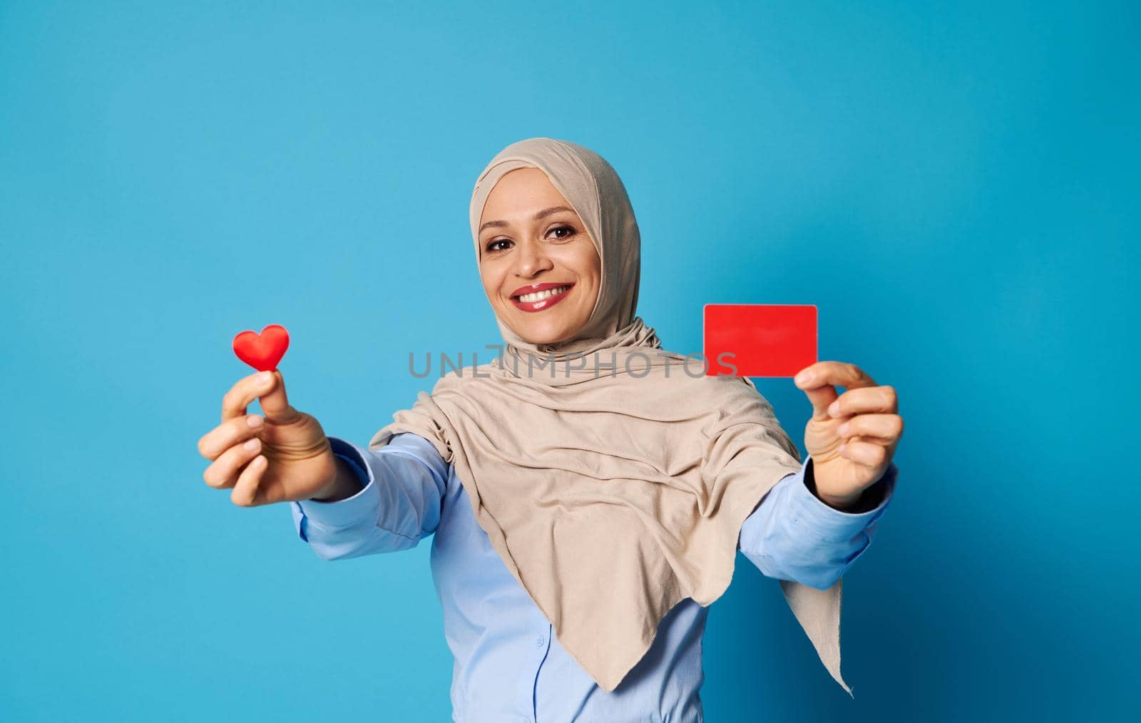 Smiling oriental woman in hijab showing a shape of red heart and blank red plastic card, smiling looking at camera while posing against blue background with copy space
