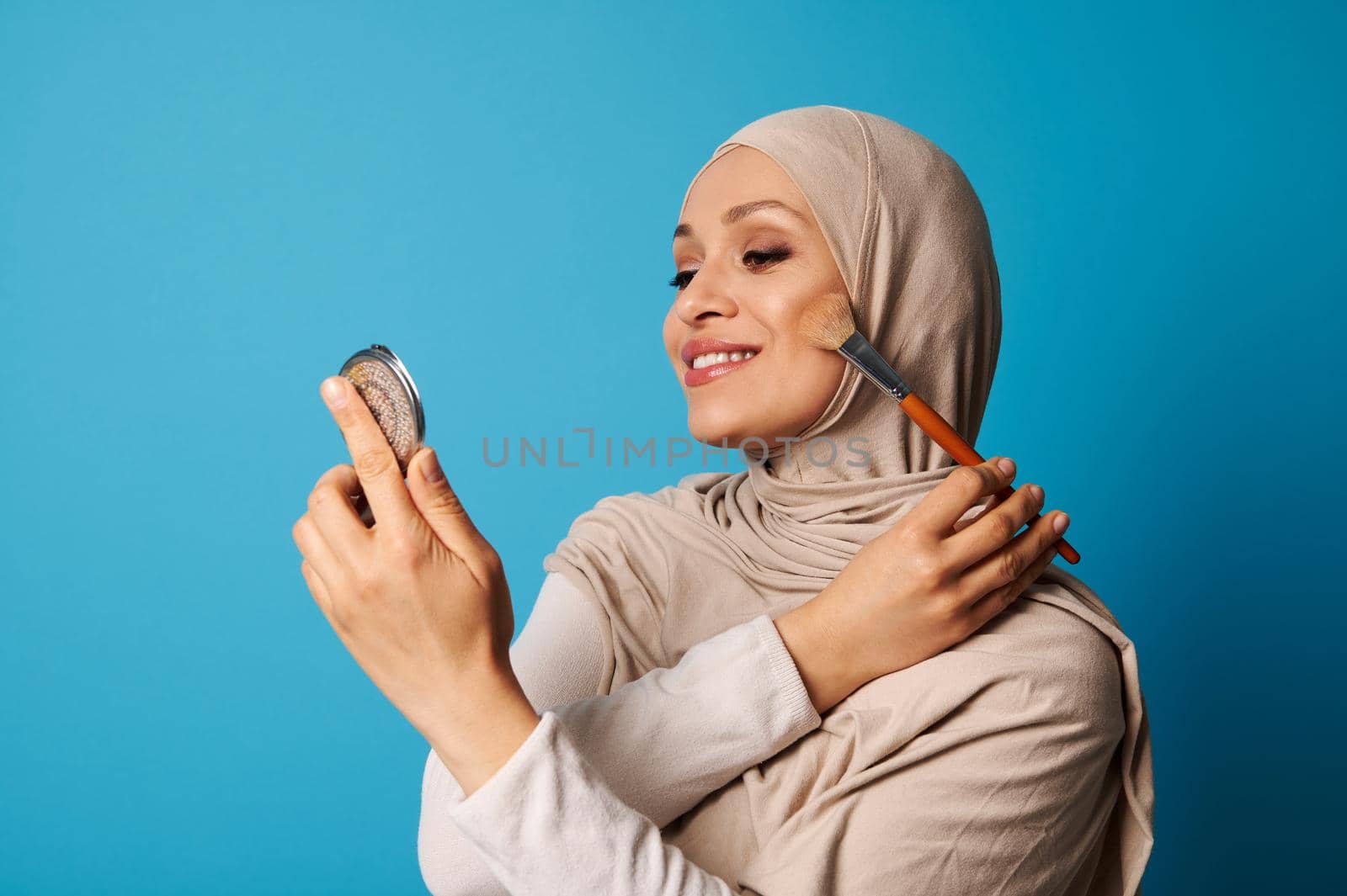 Beautiful Arabic Muslim woman in hijab holds a cosmetic mirror and a makeup brush and applies blush to the cheekbones of her face, isolated on blue background with copy space
