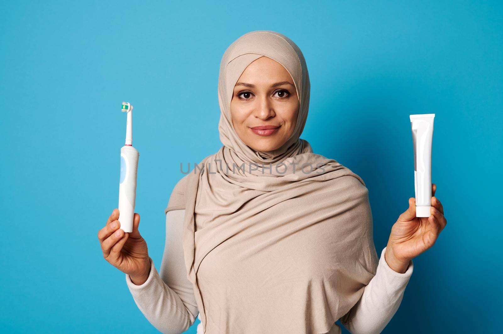 Beautiful woman with oriental appearance wearing a hijab, holding a toothpaste and toothbrush and posing looking at camera over blue background