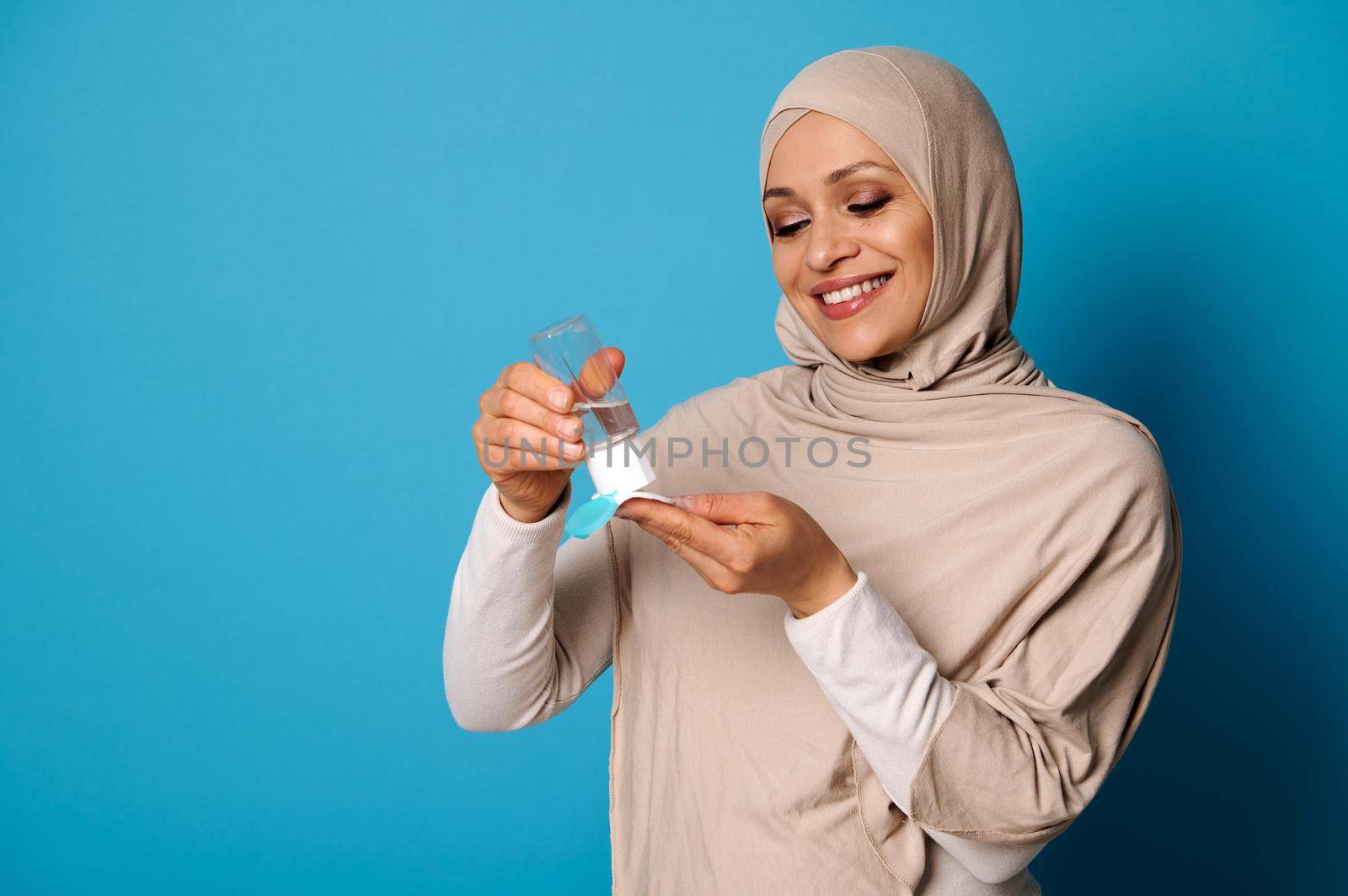 Smiling muslim woman wearing beige hijab applying micellar makeup lotion to cotton pad. Blue background, copy space by artgf