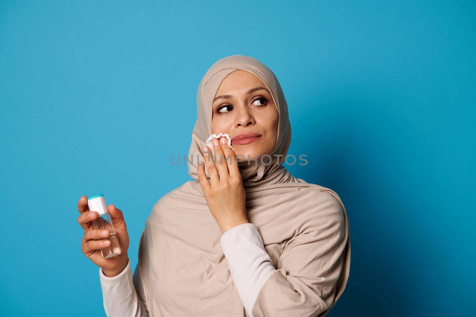 Beautiful Muslim woman with covered head in hijab applying micellar lotion and removing make up from her lips by artgf