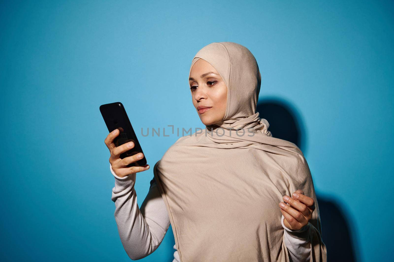 Young woman of oriental appearance in beige hijab holding a smartphone in the hand, isolated on blue background with copy space