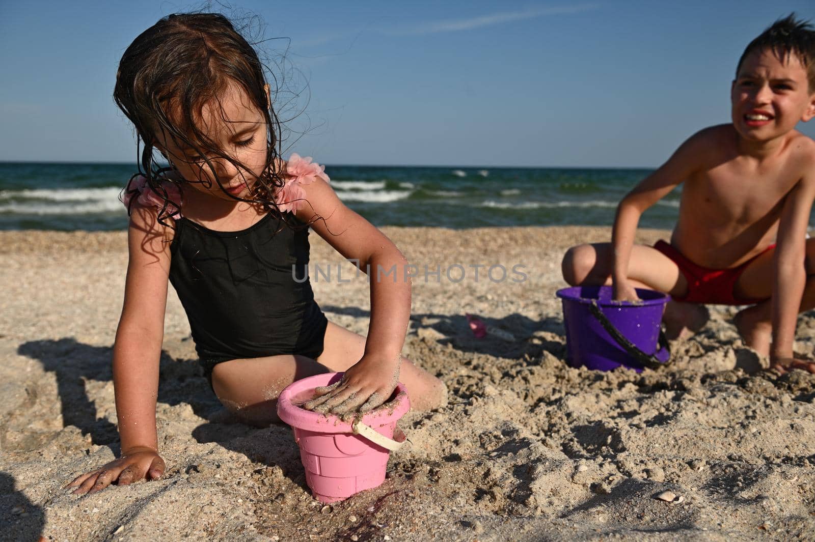 Cute girl fills a pink plastic toy bucket with sand to create figurines and builds a sand castle while playing alongside her brother against the backdrop of the sea. Summer vacation concept, fun island vacation