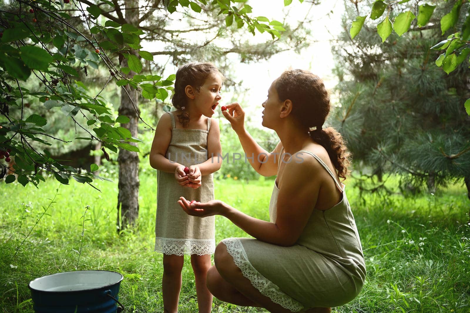 Adorable mother feeds her daughter with plucked cherries in the garden. Mom and daughter dress the same when picking cherries in summer. Motherhood, childhood, family relationships.