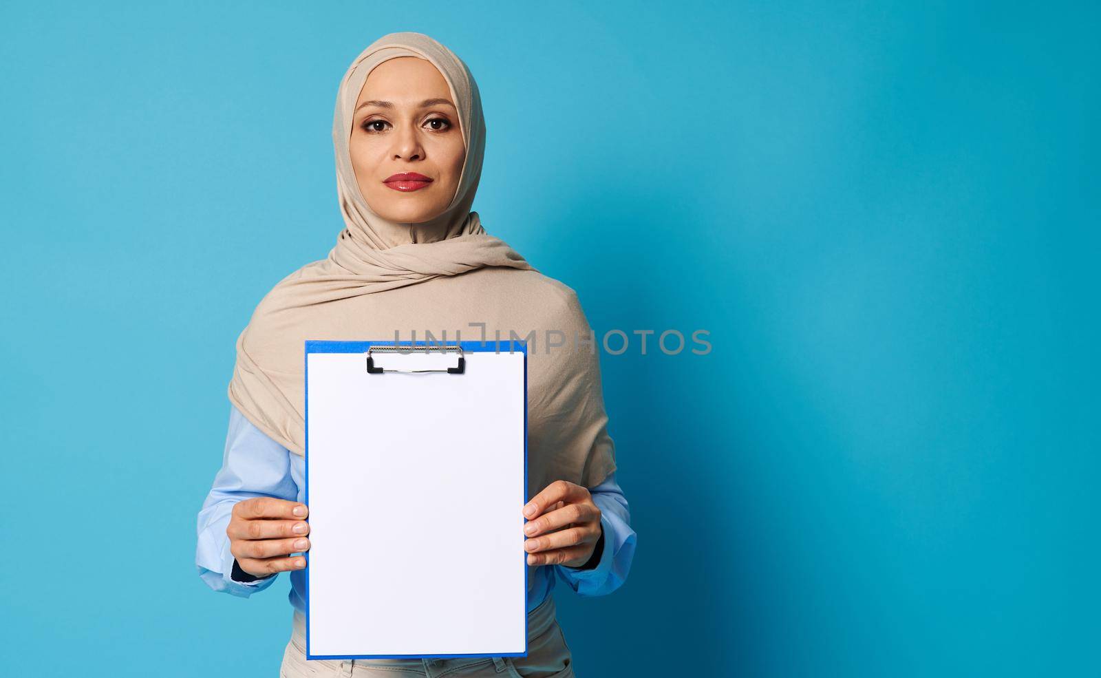 Beautiful Arab woman wearing a beige hijab holding a clipboard with white blank sheet of paper, isolated over blue background