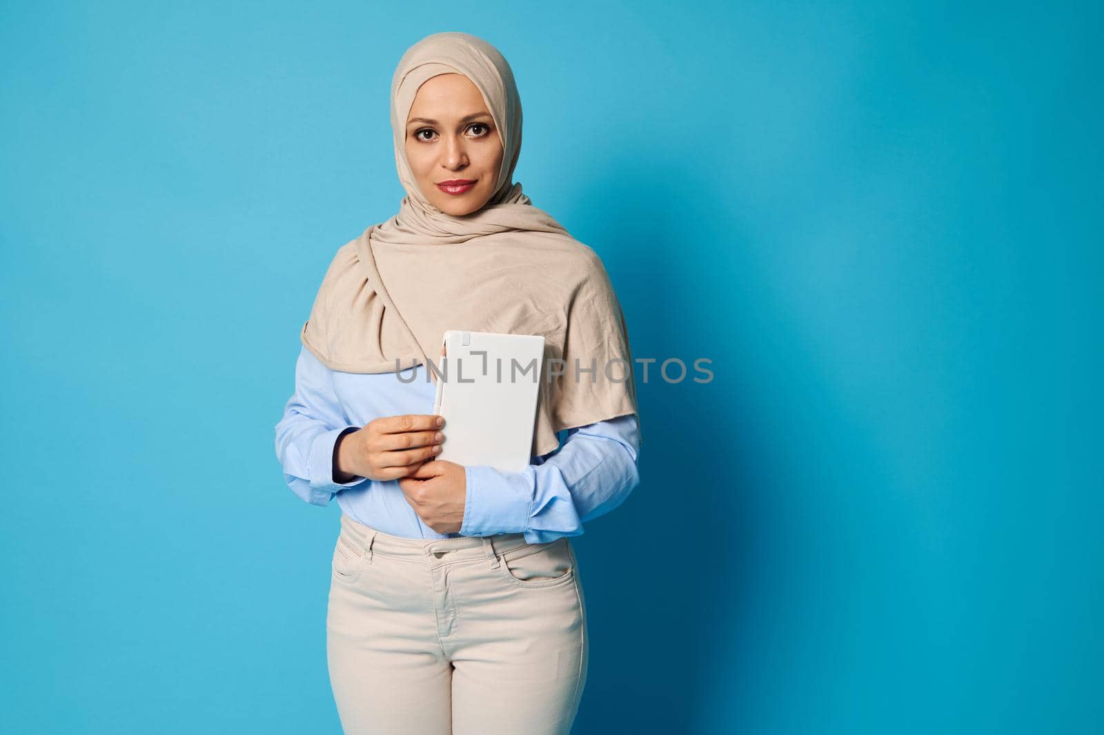 Serious and serene Arab Muslim woman with a white book in her hands. Education concepts on blue background with copy space