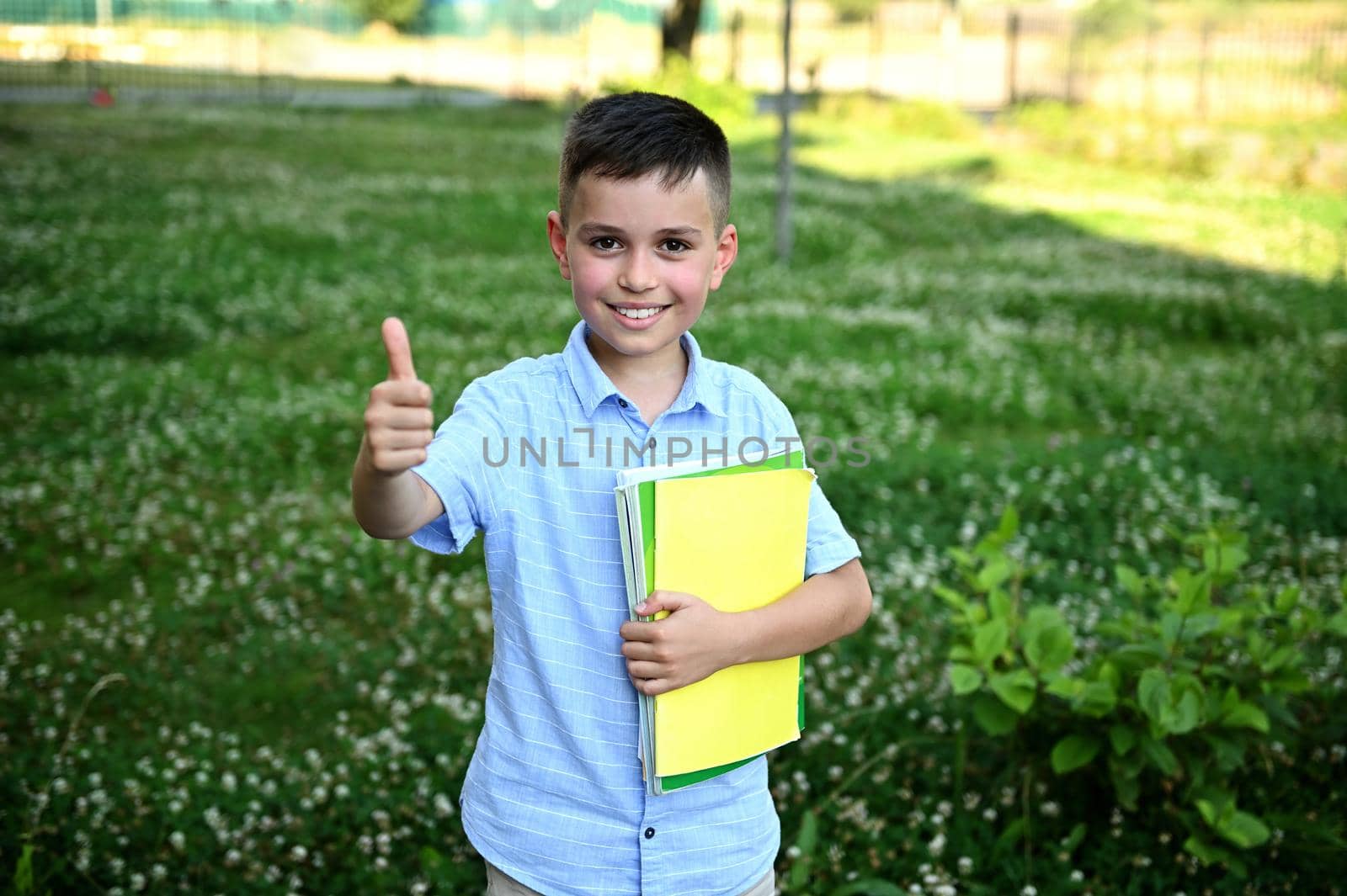 Adorable schoolboy holding a yellow workbook and showing thumb up to camera standing on green grass background by artgf