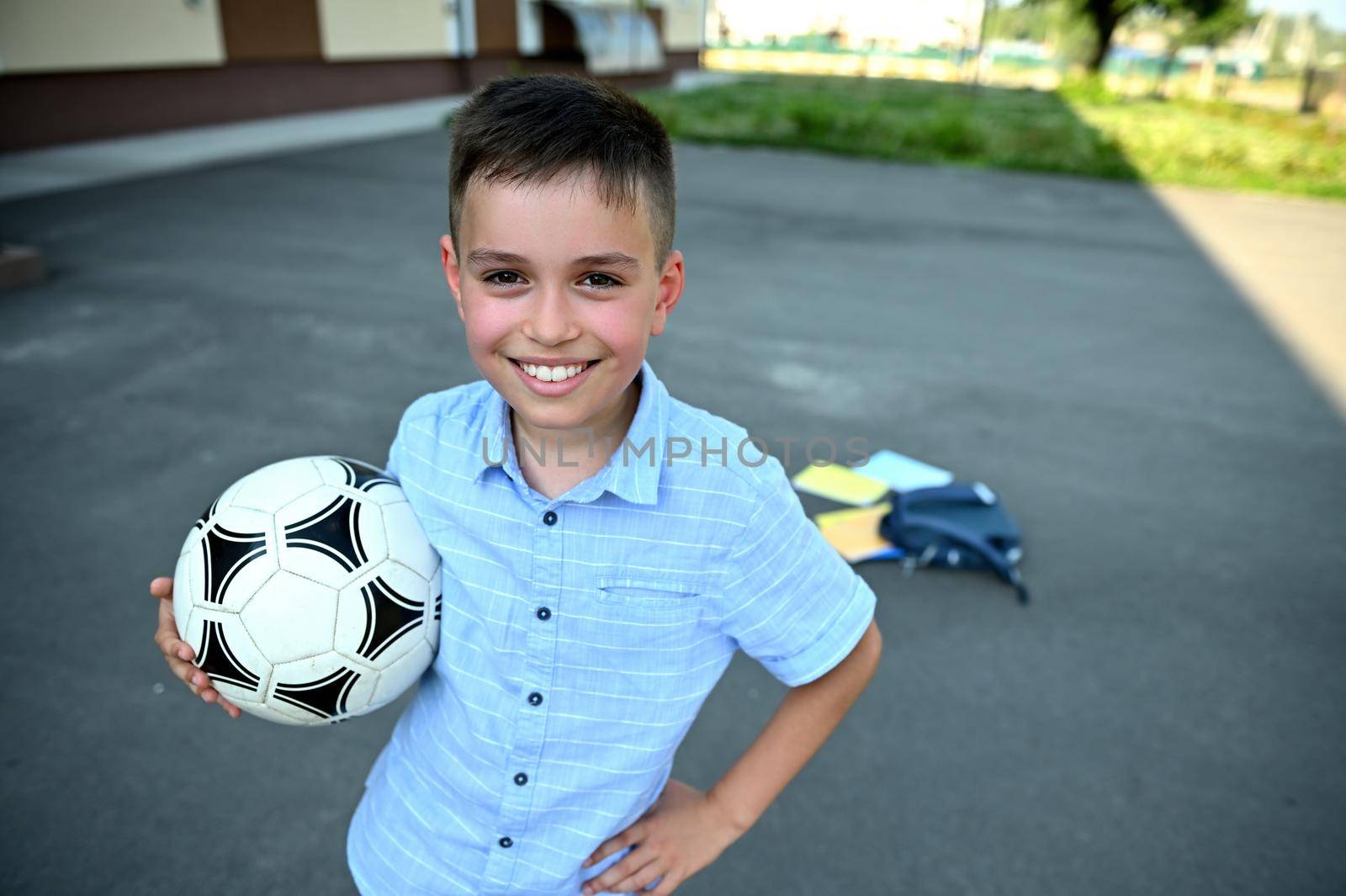 Handsome schoolboy with soccer ball in hands smiling with toothy smile on the background of scattered backpack with school supplies on the schoolyard. Recreation. Rest between lessons by artgf