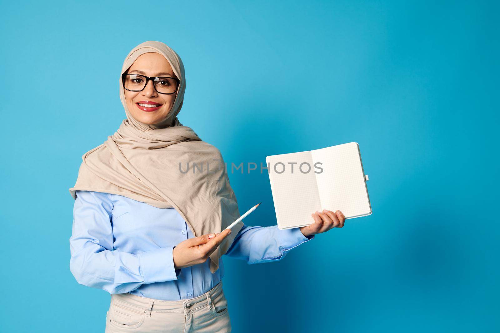 Smiling Muslim woman in hijab holds blank book and points at it with pen isolated on blue background by artgf