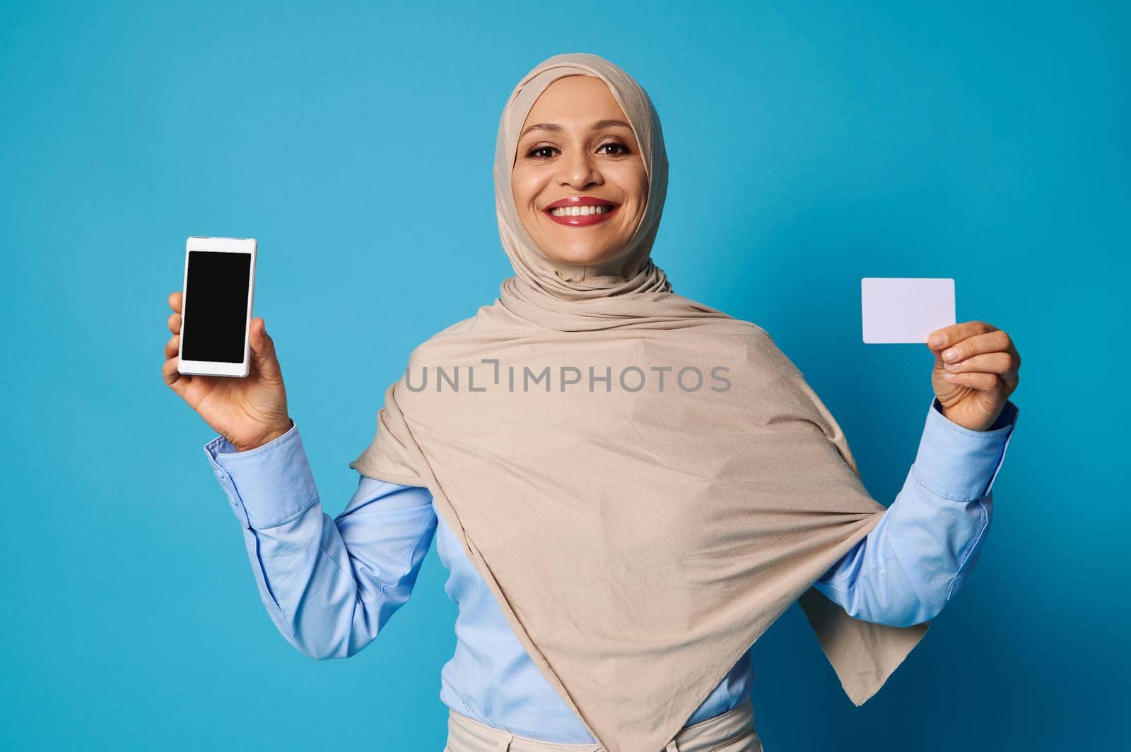 Young woman with beautiful smile holding a white blank plastic card in her hands, posing over blue background with space for text