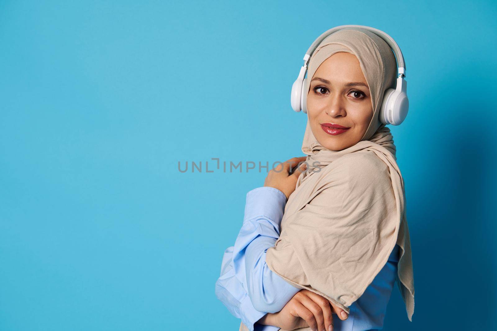 Side portrait of an attractive Oriental woman in beige hijab and headphones, posing over blue background with copy space by artgf