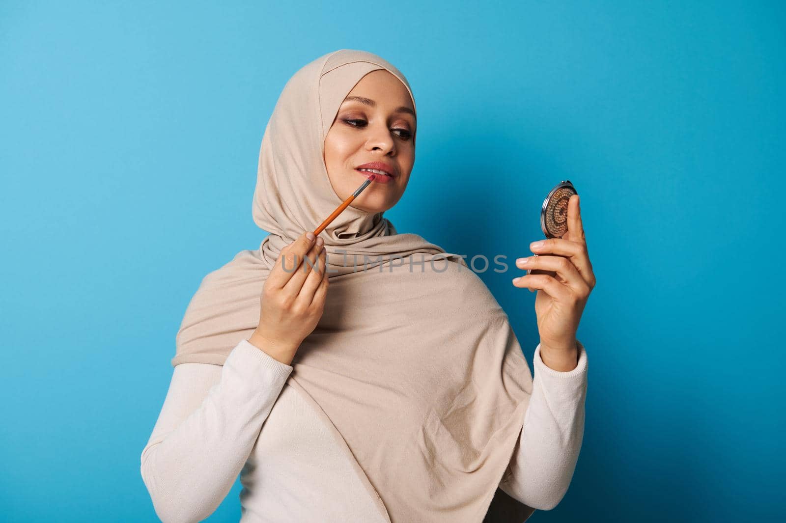 Young Arab woman in beige hijab uses a makeup brush to apply lipstick to her lips and looks at her reflection in a small cosmetic mirror. Isolated portrait on blue background with space for text