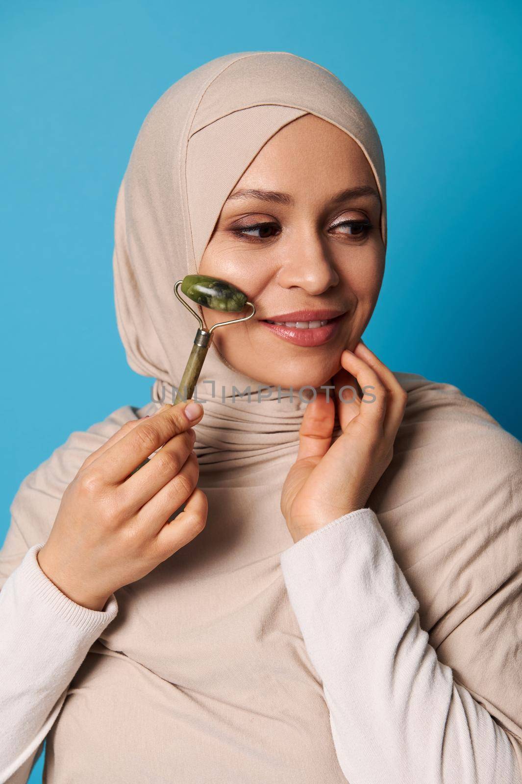 Face beauty portrait of an attractive Arabian woman in hijab and strict religious outfit using jade roller for face lymphatic drainage. Beauty treatment.