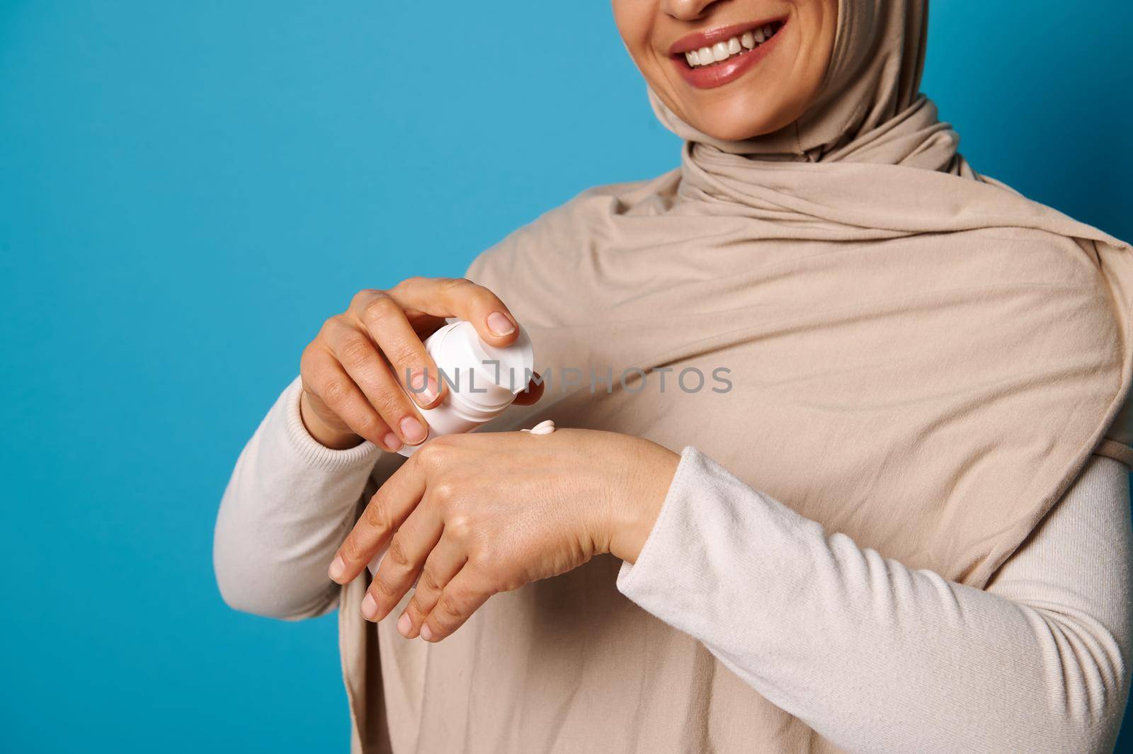 Closeup of woman in hijab squeezing cream on hand, isolated on blue background with space for text by artgf