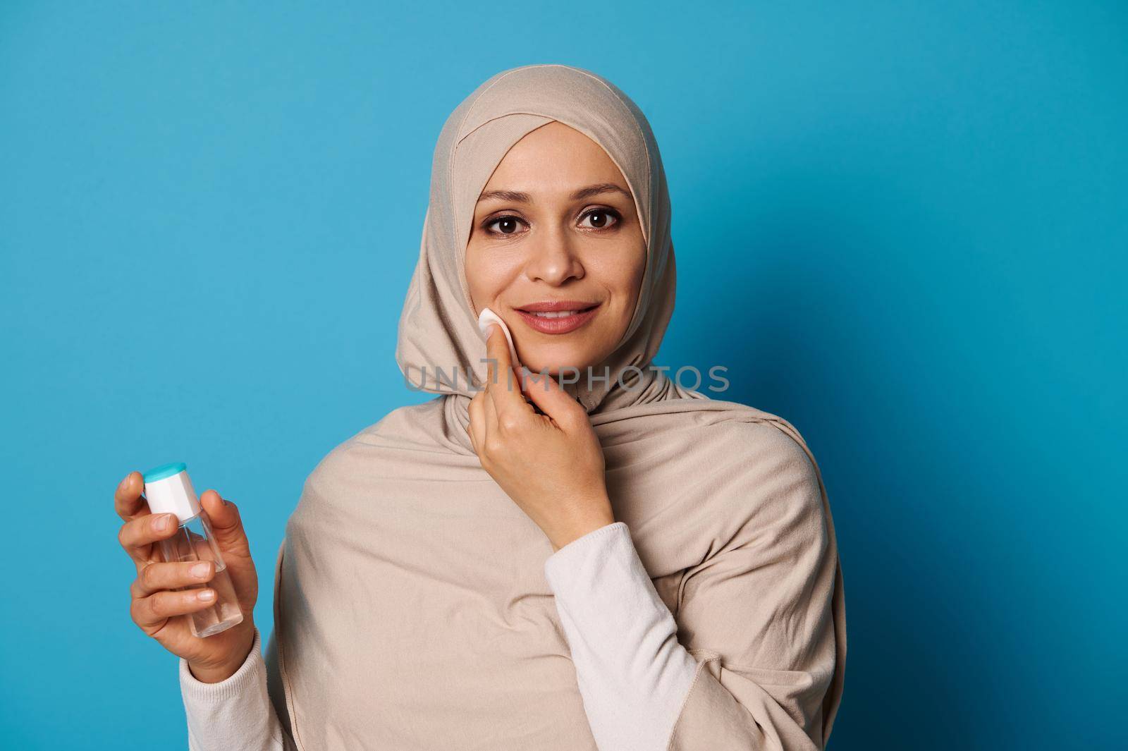 Charming Arab woman in hijab holds a cotton pad and cleans her face, smiles while looking at camera. Copy space