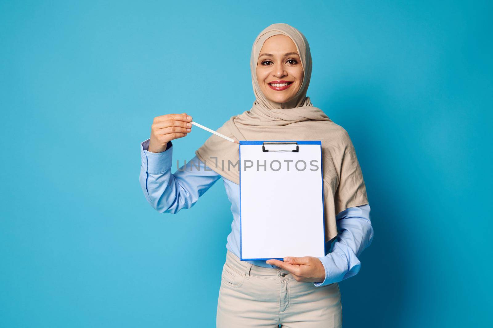 Smiling Arab woman with covered head in hijab pointing a pen on a blank surface on a white paper sheet in clipboard
