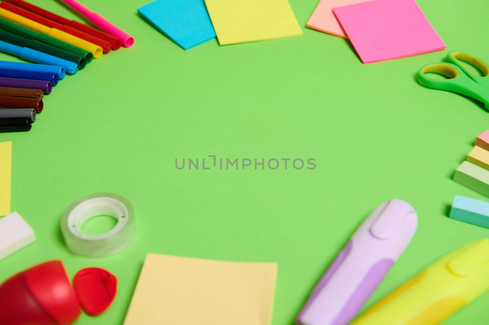 Multi colored assortment of stationery office supplies and school accessories arranged in a circle on light green background, copy space by artgf