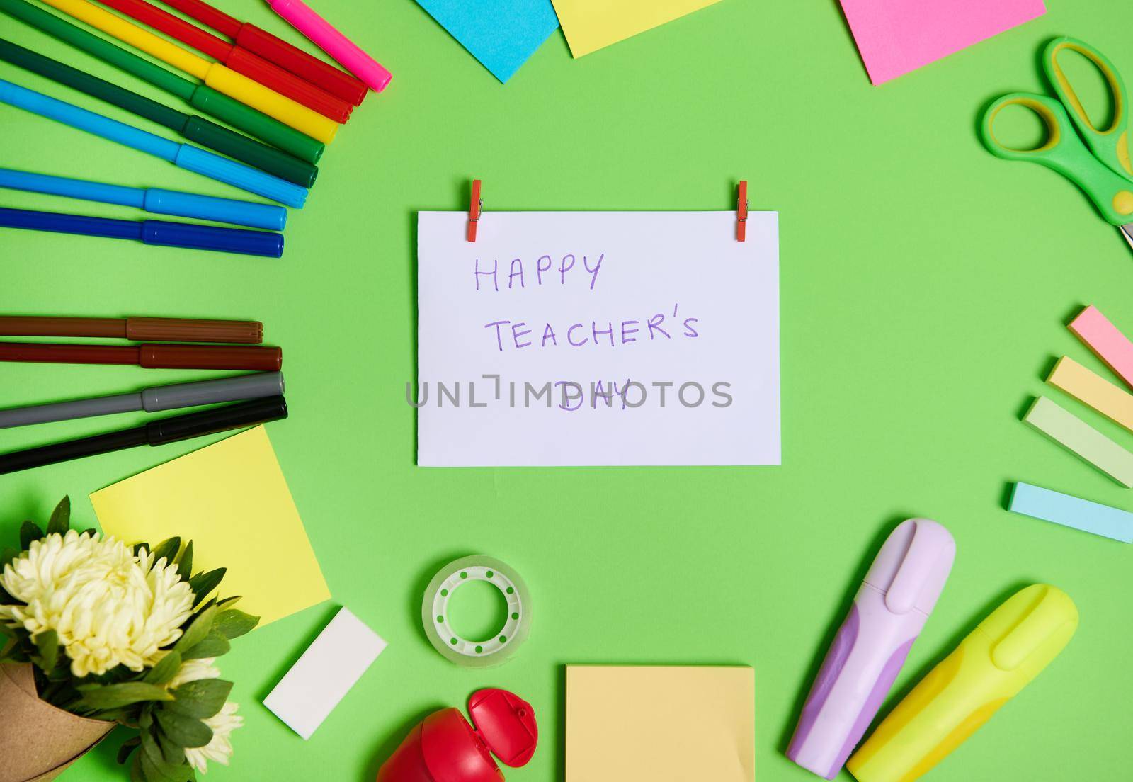 High angle view of colorful assortment of stationery office supplies and school accessories arranged in a circle, with white paper in the center with lettering Happy Teacher's Day.