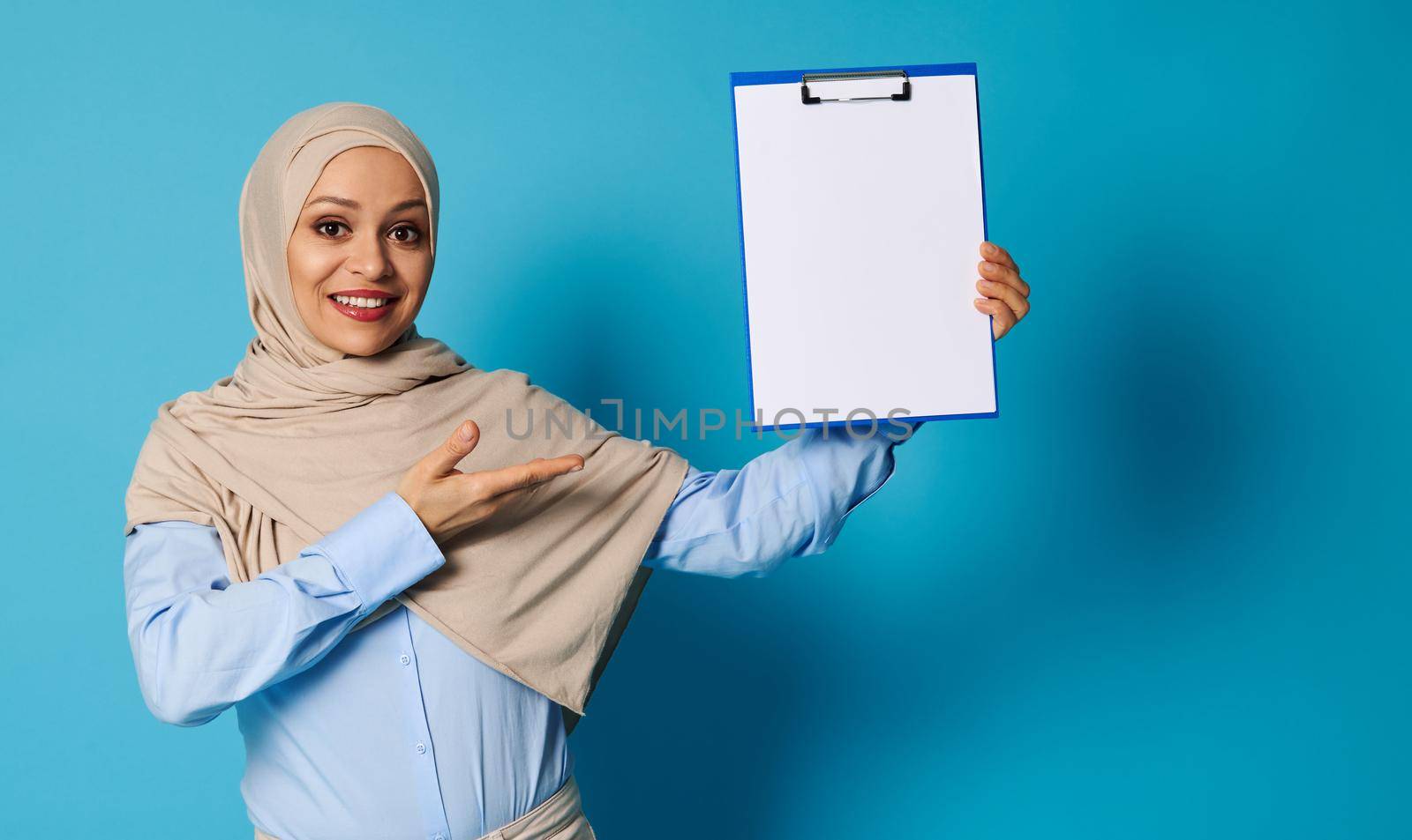 A Muslim woman with a covered head in a hijab holds a clipboard in one hand and points at it with her hands, presenting invisible information on a blank sheet of paper. Isolated over blue background