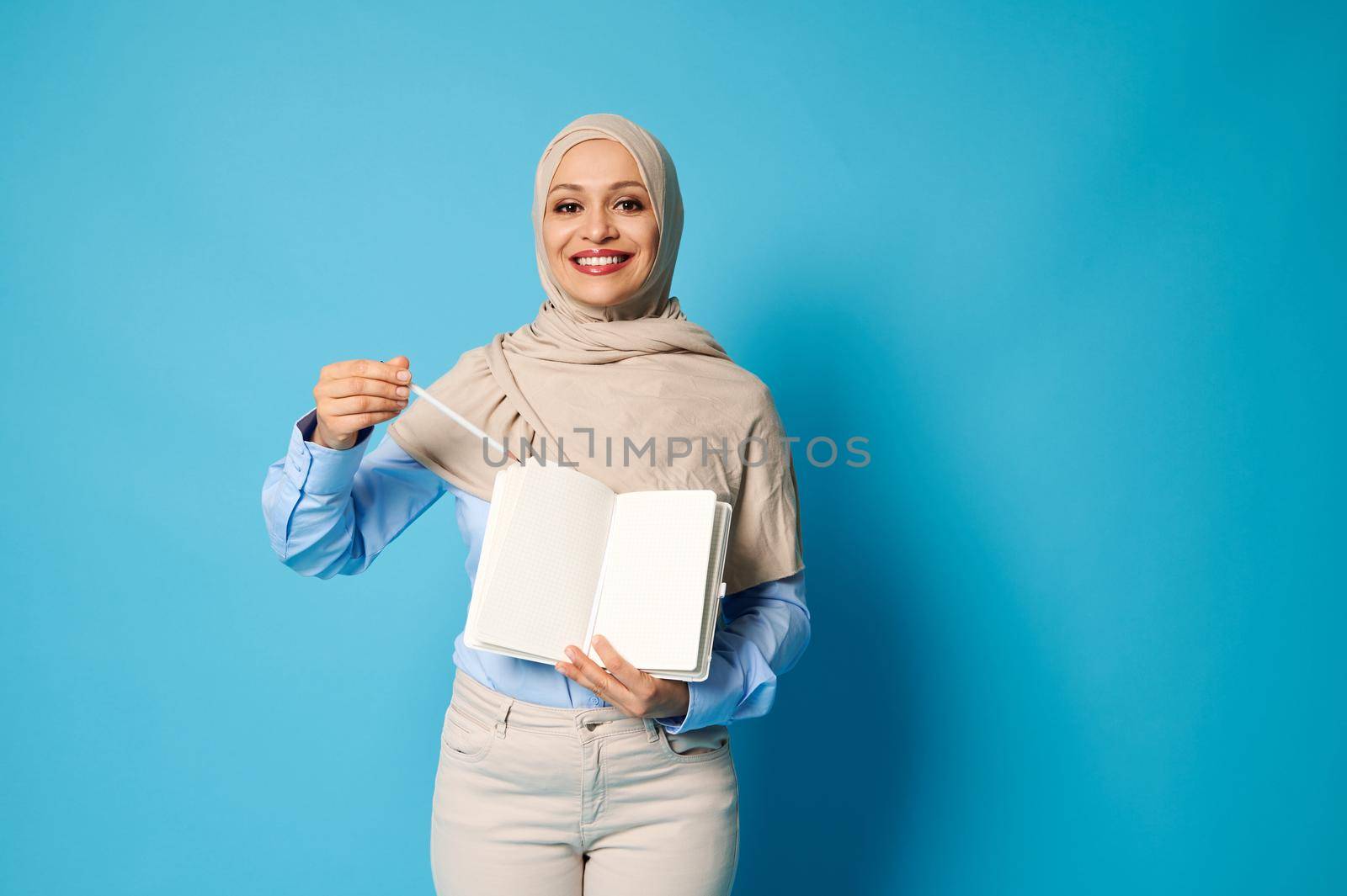 Charming Muslim woman in hijab with a beautiful smile points a pen on a white blank sheet of paper on a daily. Blue background, copy space