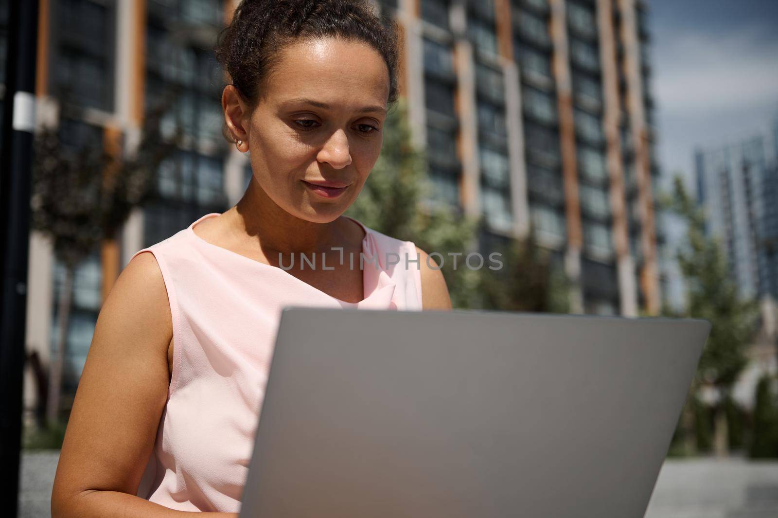 Close-up portrait of middle aged African or Latin American woman, business lady, office manager, worker, employee in casual attire working on laptop on the high buildings background