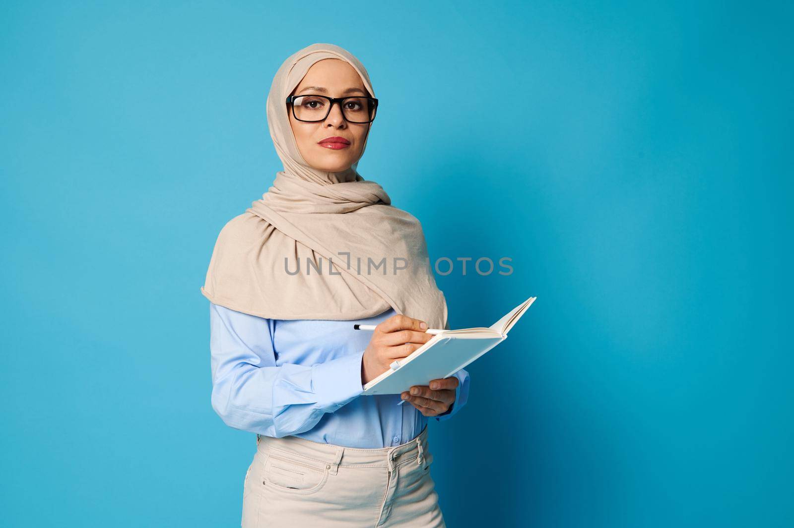 Serious Arab Muslim woman in hijab and glasses with pen and book posing on blue background with copy space