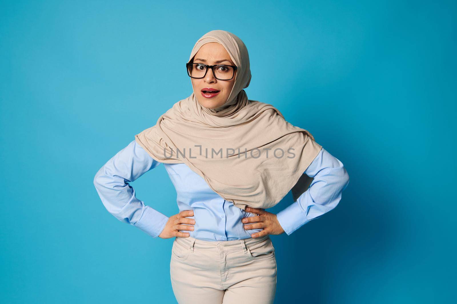 Surprised Arab Muslim woman in hijab with hands on waist expressing emotions on blue background. Concepts with copy space