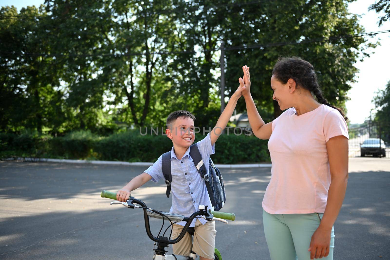 Smiling school boy on bicycle giving high five to his African American mother. Child coming back to school. Kids riding