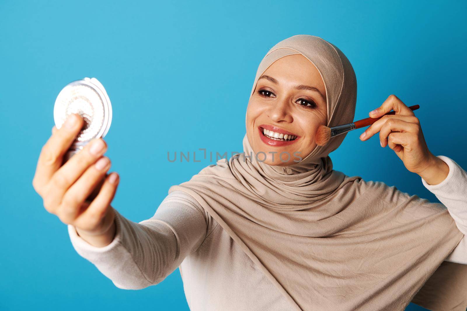 Arab Muslim woman with covered looking at mirror and applying make up, isolated on blue background with copy space