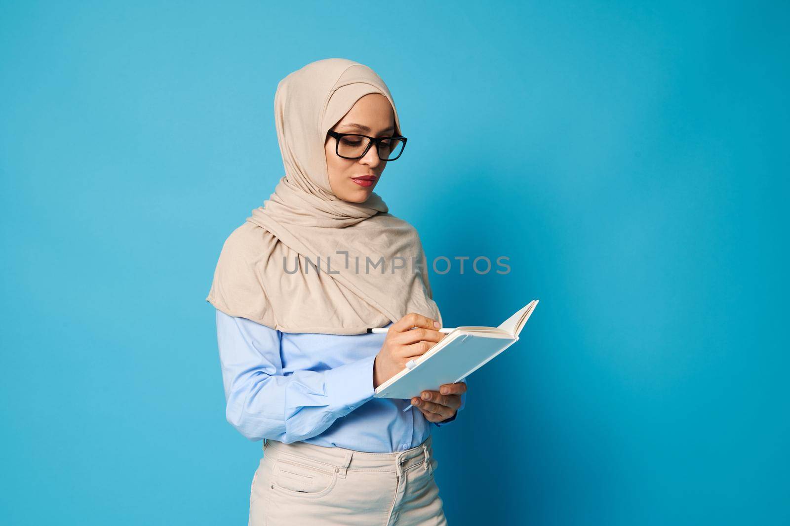 Muslim woman wearing eyeglasses and strict outfit with covered head writing on a white book, isolated over blue background with copy space