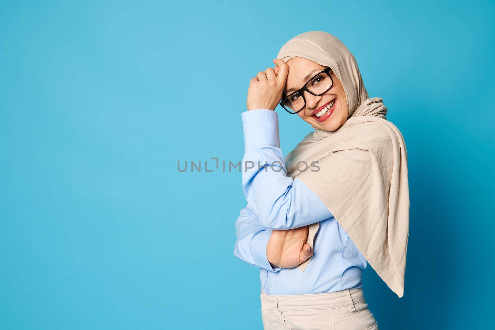 Attractive Muslim woman with covered head in beige hijab smiles with toothy smile and stands side to the camera over blue background with copy space